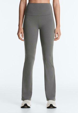 Femeie purtând pantaloni evazați, gri, mulați și cu talie înaltă, asortați cu adidași albi, stând într-un cadru alb simplu.