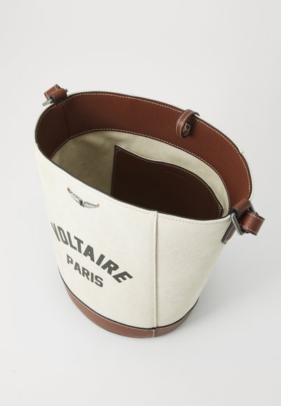 Kanvas bucket soma ar krēmkrāsas ārpusi, brūnu ādas akcentiem, ar izdrukātu "VOLTAIRE PARIS" logo un plašu brūnu iekšpusi.