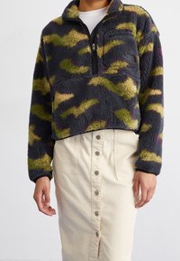 Polarowy półgolf w granatowym kolorze z zielonym i fioletowym wzorem w kamuflaż, z suwakiem na pół, wysokim kołnierzem oraz przednią kieszenią. W połączeniu z lekką spódnicą.