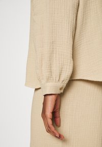 Beige, strukturovaný top s dlouhým rukávem a nařaseným manžetou s detaily knoflíků. Tkanina se jeví jako jemná a lehká, což zdůrazňuje uvolněný střih.