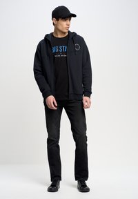 Sudadera negra con cremallera y bolsillos frontales, camiseta negra con texto azul, jeans negros y zapatillas negras. Gorra puesta al revés.
