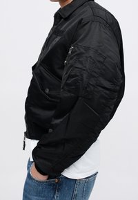 Personne portant une veste bomber noire avec des poches zippées et à boutons-pression, un jean bleu et une chemise blanche visible à la taille.