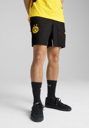 Melni sporta šorti ar dzelteno BVB logo, ar gludu audumu un divām baltām sānu svītrām, apvienoti ar melnajām zeķēm un sporta apaviem.