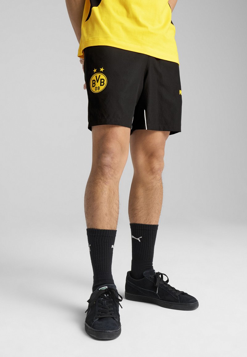 Sorte træningsshorts med gult BVB-logo, der har et glat stof og to hvide sidelæg, parret med sorte sokker og sneakers.