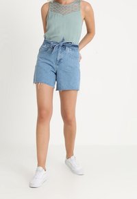 Ljusblå denimshorts med hög midja, fransad fåll och ett matchande bälte. Kombineras med en ärmlös mintgrön topp med spetsdetaljer.