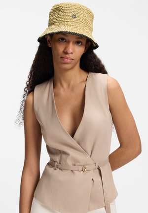 Mujer con cabello largo y rizado, lleva un top beige sin mangas con cinturón y un sombrero tipo pescador tejido color canela con ribete negro.