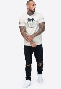 Kratki kremasti majica s kratkim rukavima s crnim grafičkim prikazom lava i natpisom "Lonsdale London", uparen s izblijedjelim crnim trapericama i bijelim tenisicama.