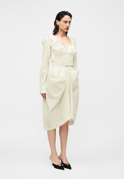 Γυναίκα που φορά ένα εκρού midi φόρεμα με ζώνη, μακριά μανίκια, κυματιστό ντεκολτέ και δύο μεγάλες μπροστινές τσέπες, φορώντας μαύρες μυτερές γόβες.