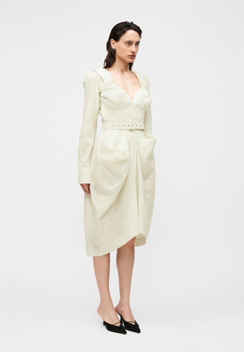 Femme debout dans une robe midi écrue ceinturée avec manches longues, encolure festonnée et deux grandes poches avant, portant des escarpins noirs pointus.