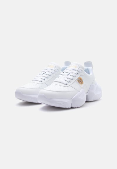Paire de baskets blanches épaisses avec semelles épaisses et lacets à l'avant, présentant un petit emblème doré et blanc sur les panneaux latéraux.
