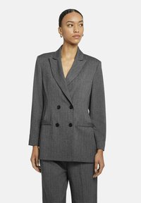 Grijze dubbelzijdige blazer met een gestructureerde afwerking, klassieke revers, twee voorzakken en zwarte knopen. Gecombineerd met bijpassende broeken.