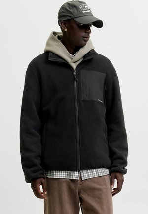 Homme portant une veste polaire noire zippée sur un sweat à capuche vert et une chemise à carreaux, pantalon marron, et une casquette grise avec un logo, regardant sur le côté.
