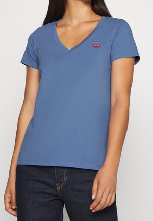 Kobieta w niebieskim T-shircie Levi's z dekoltem w serek, krótkimi rękawami i ciemnoniebieskich dżinsach na jednolitym tle.