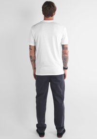 Homme avec des bras tatoués portant un t-shirt blanc uni, un pantalon sombre et des chaussures noires, se tenant de dos contre un fond blanc.