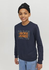 Mornarsko plavi dukserica sivoj pravokutnoj grafici s natpisom "JACK&JONES" u narančastoj boji, rebrasti izrez na vratu i manžetama. Nosio se uz traperice.