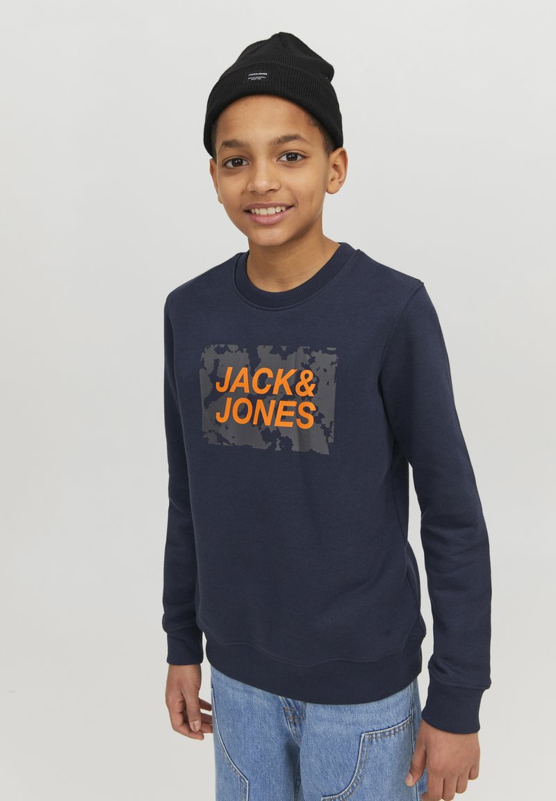 Mornarsko plavi dukserica sivoj pravokutnoj grafici s natpisom "JACK&JONES" u narančastoj boji, rebrasti izrez na vratu i manžetama. Nosio se uz traperice.