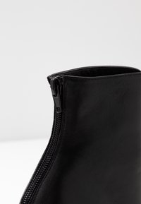 Détail d'une botte noire en cuir arrivant à la cheville montrant une fermeture éclair noire fermée sur le côté, sur un fond blanc uni.