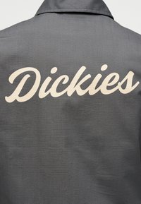 Γκρι ύφασμα με μεγάλο λευκό λογότυπο "Dickies" σε πλάγια γραφή τυπωμένο στην πίσω πλευρά ενός ένδυματος με γιακά.
