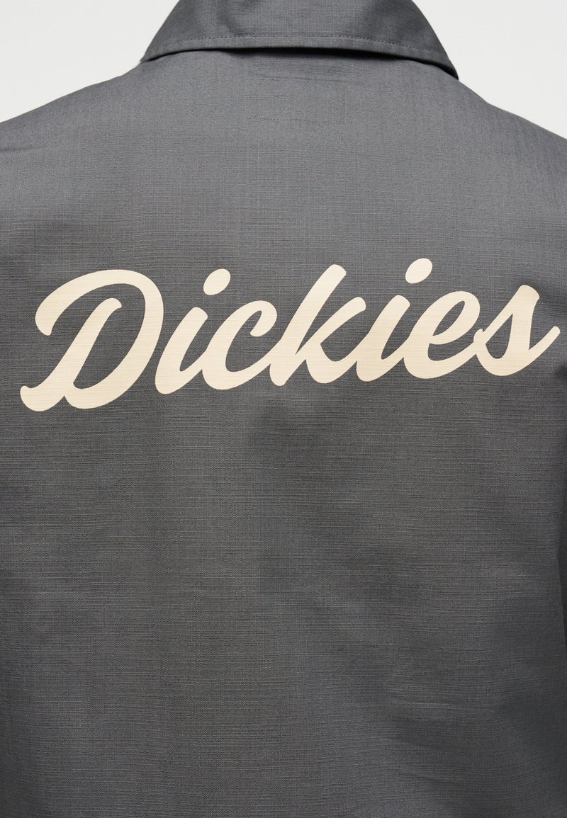 Grauer Stoff mit großem weißem kursivem "Dickies"-Logo auf der Rückseite eines Kleidungsstücks mit Kragen bedruckt.