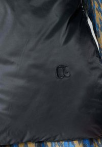 Pochette en tissu noir avec une texture lisse, ornée d'un logo brodé subtil. L'arrière-plan présente un tissu à motifs en bleu et beige.