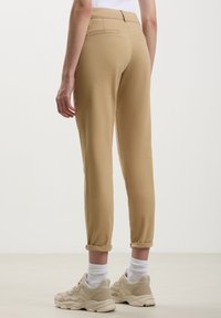 Pantaloni beige a gamba affusolata con una superficie liscia, con risvolti e una tasca posteriore. Abbinati a sneakers di colore chiaro e calzini bianchi.