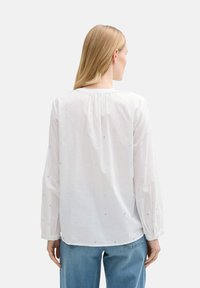 Chemise blanche à manches longues avec des points brodés multicolores, détails froncés au dos et poignets boutonnés. Portée avec un jean bleu.