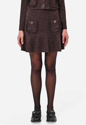 Donna con gonna marrone in tweed con due tasche frontali, collant neri trasparenti e mocassini neri borchiati su sfondo bianco.