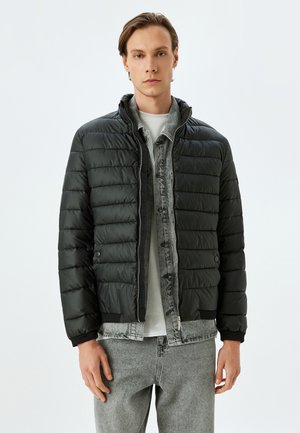 Czarna pikowana kurtka puchowa z zamkiem błyskawicznym i wysokim kołnierzem, o pikowanej teksturze i ściągaczach przy rękawach, noszona na szarej koszuli jeansowej.