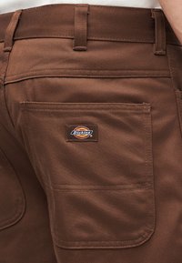 Καφέ εργασιακό παντελόνι με πίσω τσέπη και εμφανές λογότυπο Dickies, που φοριέται από άτομο με λευκό πουκάμισο.