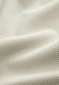 Texturovaná tkanina světle béžové barvy s mřížkovým vzorem, obsahující měkká tkaná vlákna, která vytvářejí jemné vlny a záhyby.