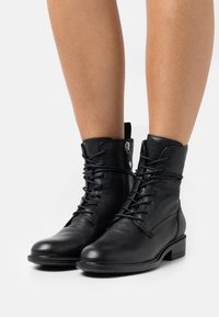 Botas de cuero negro hasta el tobillo con cordones en la parte delantera, punta redondeada y tacón bajo en bloque. Incluyen una cremallera lateral y detalles texturizados en la superficie.
