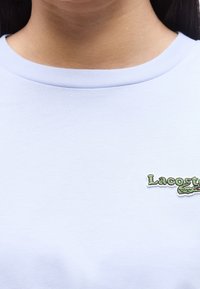 Gros plan d’une personne portant un t-shirt bleu clair avec un petit logo vert Lacoste sur le côté droit de la poitrine.