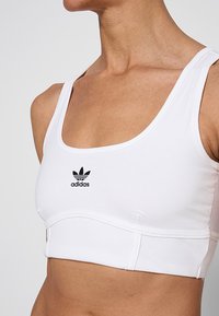 Baltas sportinis trumpas viršus, pagamintas iš tamprios medžiagos, su kvadratine iškirpte ir juodu Adidas logotipu priekyje, su riboto dizaino apatine juosta.