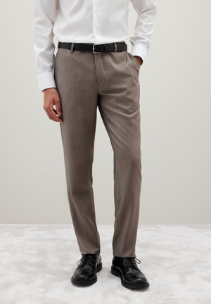 Pantalones marrones de un tejido texturizado, con parte frontal plana, trabillas para cinturón y bolsillos laterales, combinados con zapatos de cuero negros.