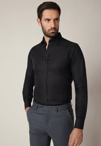 Czarna, dopasowana koszula z długim rękawem, zapinana na guziki, z prosto wyciętym kołnierzem. Połączona z szarymi, dopasowanymi spodniami.