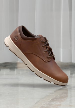 Brown leather shoe with a rounded toe, contrasting cream sole, and brown accents. Features lacing and a logo on the side.