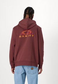 Bordeaux színű kapucnis pulóver, nagy, narancs és piros "Oakley" grafikával és kör alakú mintával a hátsó részén. Puha pamut anyagból készült, laza szabással.