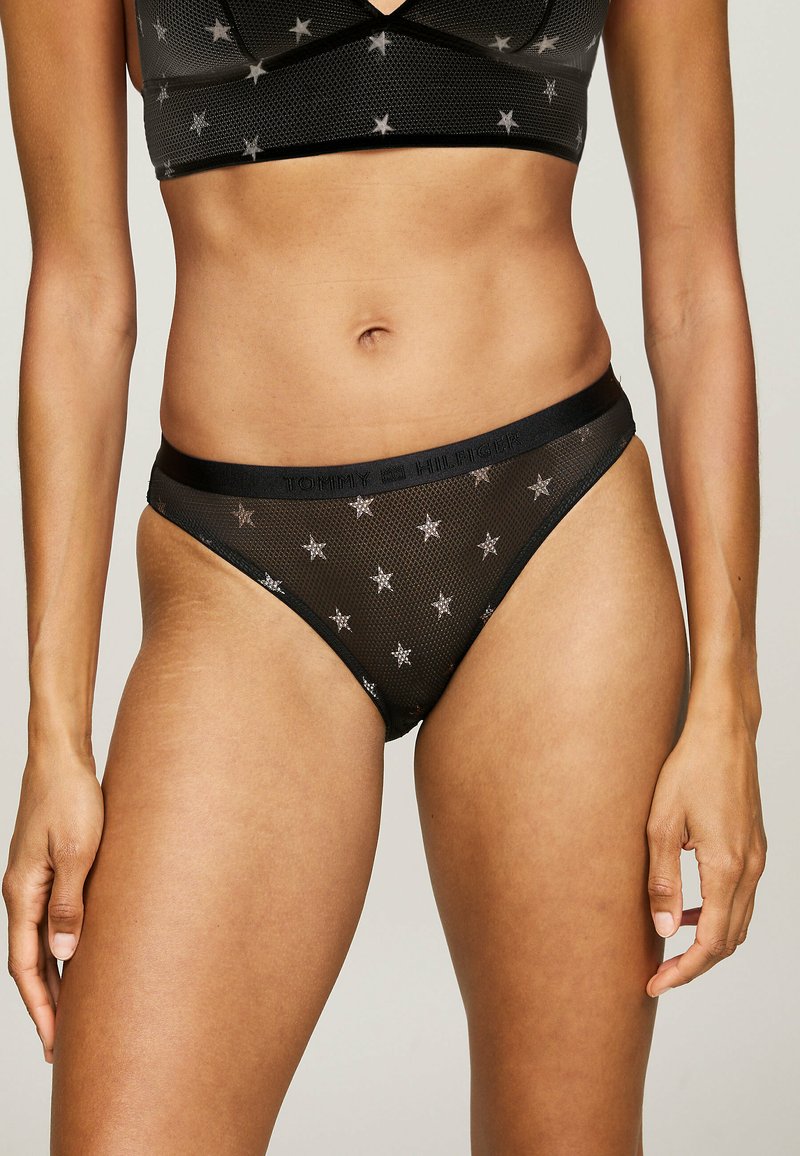 Bralet din plasă neagră cu model de stele argintii și un thong din plasă neagră asortat, având un brâu cu marcă și o textură netedă.