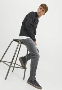 Jachetă din denim negru peste un tricou alb, asortată cu blugi gri și adidași bleumarin. Așezat pe un taburet metalic, cu fața ușor întoarsă.