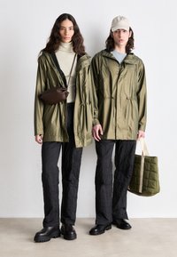 Two individuals wearing olive green rain jackets, black padded pants, and black chunky boots. One holds a quilted olive bag, the other a brown crossbody.