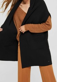 Zwarte open voorvest met twee voorzakken, gedragen over een roestkleurige, gestructureerde brei trui en tankleurige broek. Zachte stof en een relaxte pasvorm.