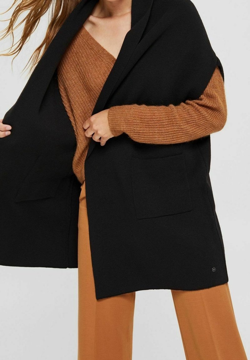 Zwarte open voorvest met twee voorzakken, gedragen over een roestkleurige, gestructureerde brei trui en tankleurige broek. Zachte stof en een relaxte pasvorm.