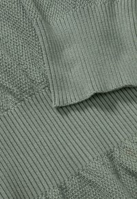 Dettaglio di un maglione verde lavorato a maglia con un motivo testurizzato, orlo a coste e sezioni lisce, che mostra un tessuto morbido e caldo.