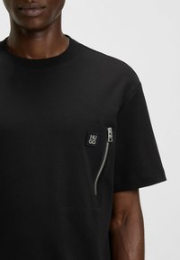 T-shirt nera con vestibilità rilassata, dotata di una tasca con zip sul lato sinistro del petto e un patch con logo. Realizzata in morbido cotone e con maniche corte.
