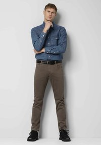 Chemise en denim à boutons et deux poches poitrine, associée à un pantalon slim-fit marron et des baskets noires, sur un fond neutre.