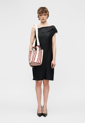 Γυναίκα με μαύρο ασύμμετρο φόρεμα που κρατά μια λευκή τσάντα ώμου με κόκκινες ρίγες, φοράει μαύρες γόβες, όρθια.