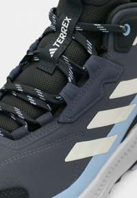 Σκούρο γκρι παπούτσι πεζοπορίας με υφή υφάσματος, μπλε λεπτομέρειες και άσπρες ρίγες. Διαθέτει αναπνεύσιμο επάνω μέρος από δίχτυ και ενισχυμένες θηλιές.