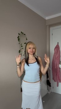 Jeune femme aux cheveux blonds et noirs porte un haut bleu clair et une jupe blanche, sourit les mains levées, debout dans une chambre bien rangée.