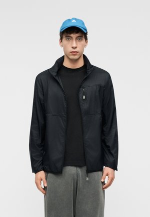 Mladý muž v modrej čiapke, čiernej bunde na zips a voľných sivých nohaviciach stojí na jednoduchom svetlom pozadí.