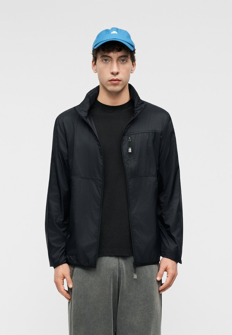 Jeune homme portant une casquette bleue, une veste zippée noire sur un t-shirt noir, et un pantalon gris ample, debout devant un fond clair uni.